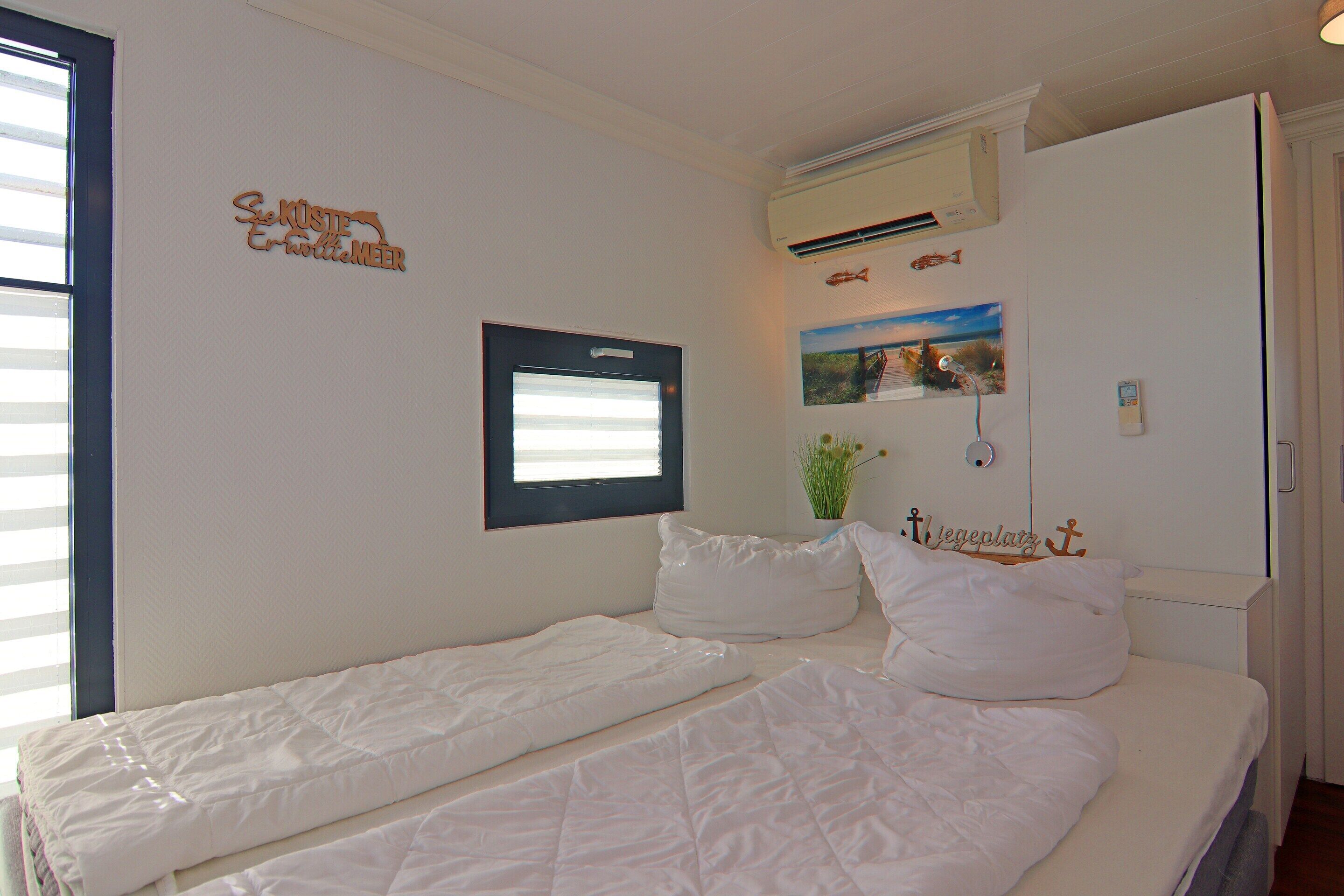 Ein Schlafzimmer mit zwei Betten, einem Fenster, einer Wandklimaanlage und Dekorationselementen, darunter ein gerahmtes Bild und ein Schild.