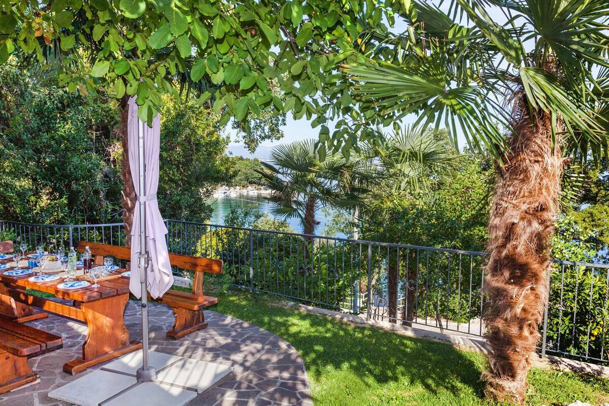 An outdoor dining area with a stone patio, wooden tables, and benches, surrounded by lush greenery and a view of a body of water.