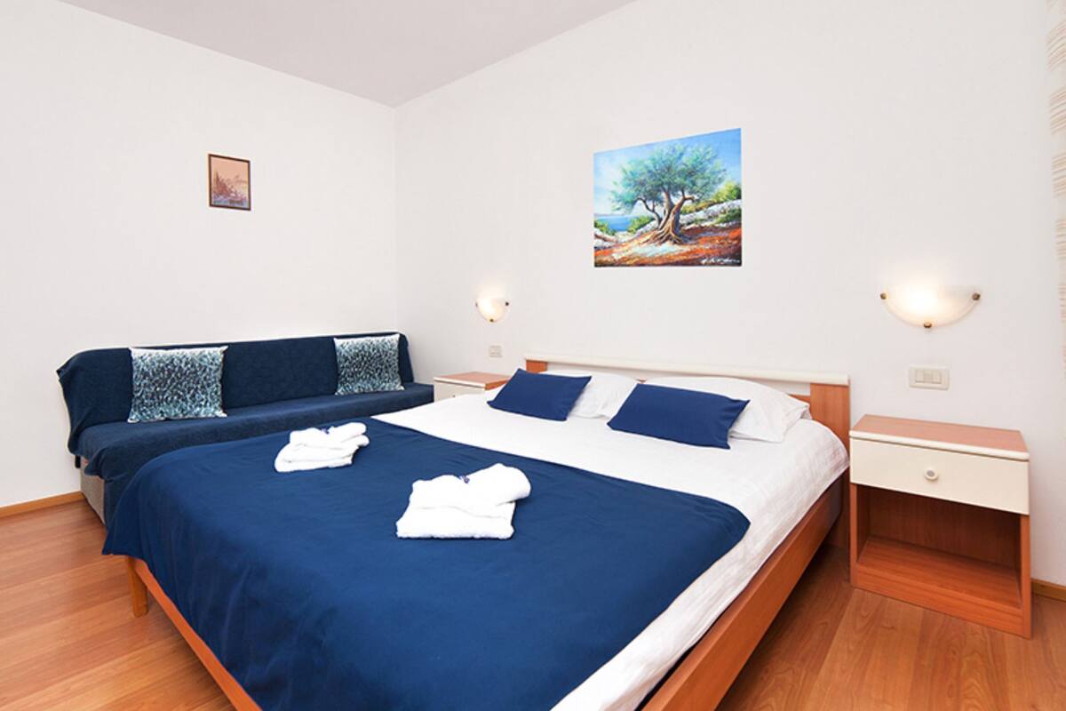 A double bed with blue and white bedding, a bedside table with a lamp, and a painting of a tree on the wall.