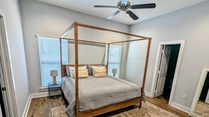 A four-poster bed with a grey upholstered headboard, a patterned rug, and a ceiling fan.