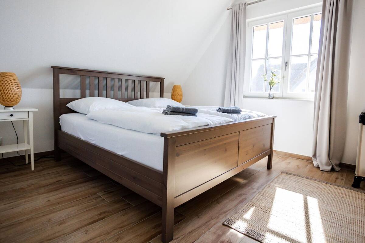 A neatly made bed in a room with wooden flooring, a bedside table with a lamp, and a window with curtains.