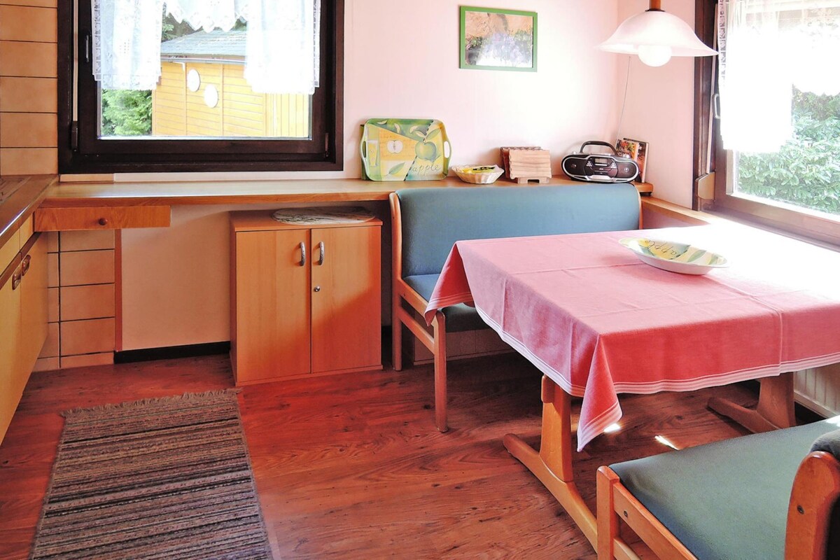 Ett mysigt rum med ett skrivbord i trä, en gungstol, ett bord med en rosa duk och ett fönster med utsikt över ett hus.