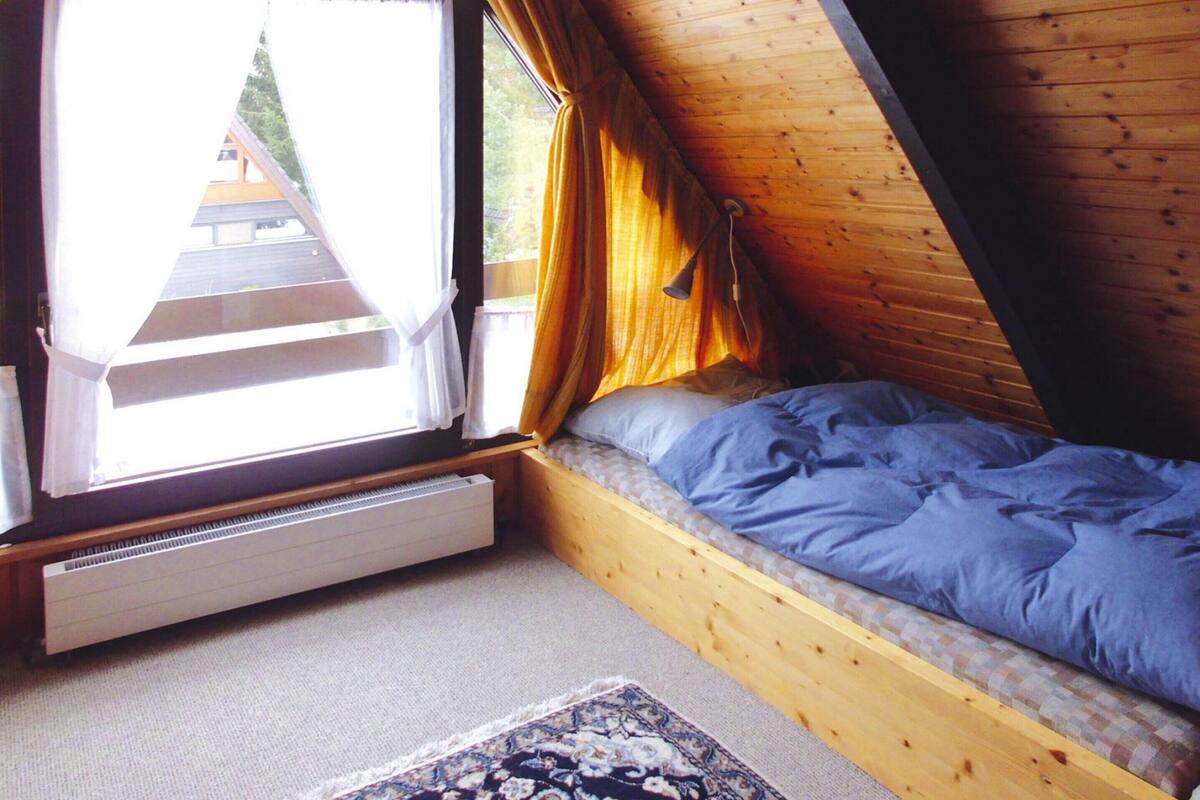 Ett sovrum med ett sluttande tak, en säng med blått sänglinne, ett element och ett fönster med gardiner.