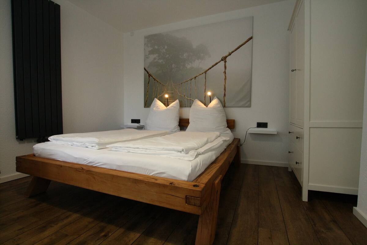 Un lit en bois aux allures soignées, agrémenté de draps blancs, une table de chevet, une petite commode et une lampe fixée au mur.