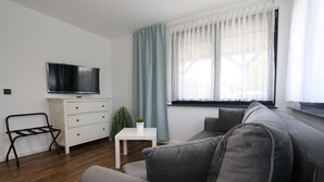 En moderne stue med en grå sofa, et hvidt sofabord, et fjernsyn på et hvidt skab og et vindue med lette gardiner.