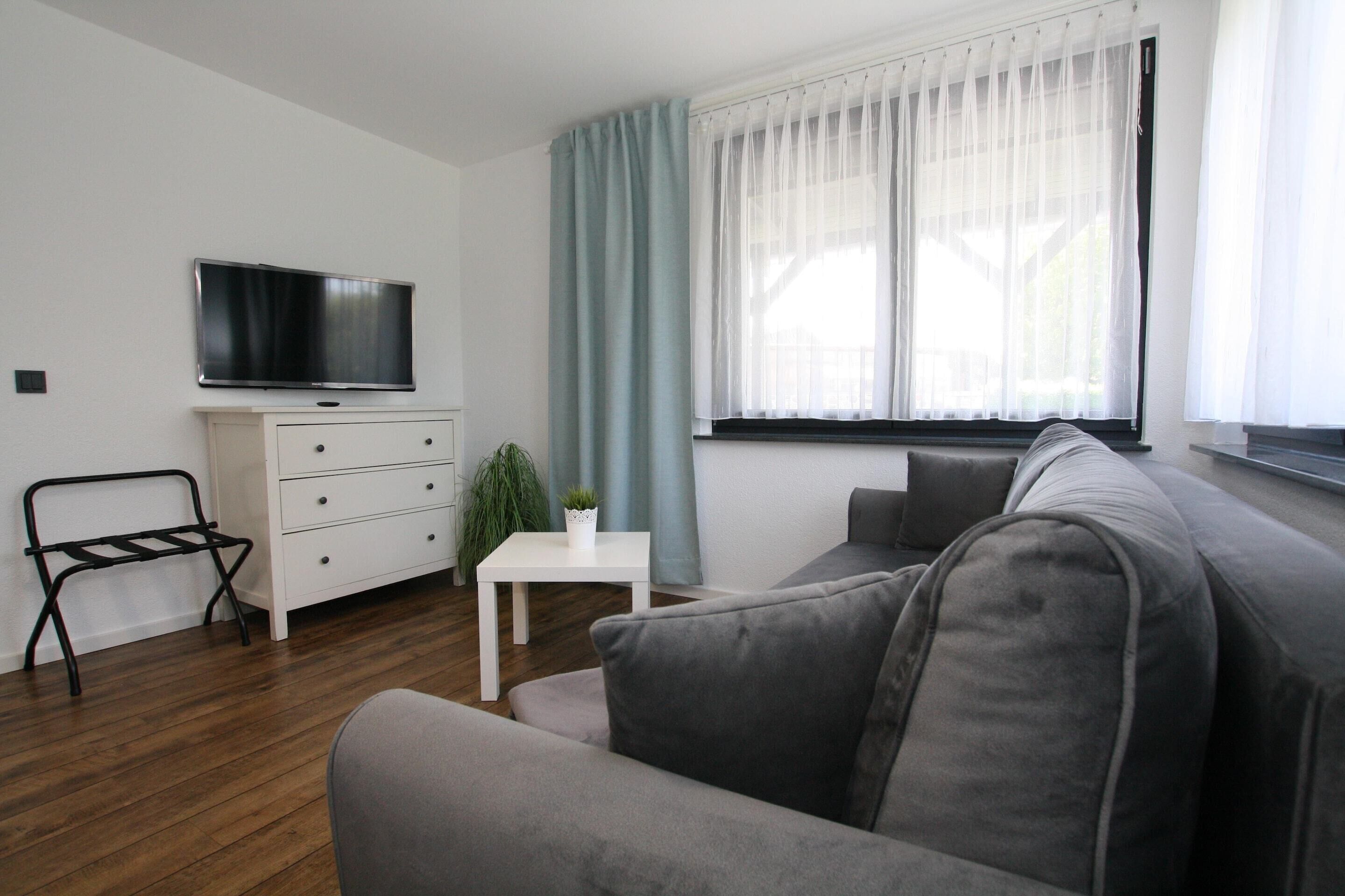 A modern living room with a grey sofa, a white table, a TV on a white cabinet, and a window with sheer curtains.