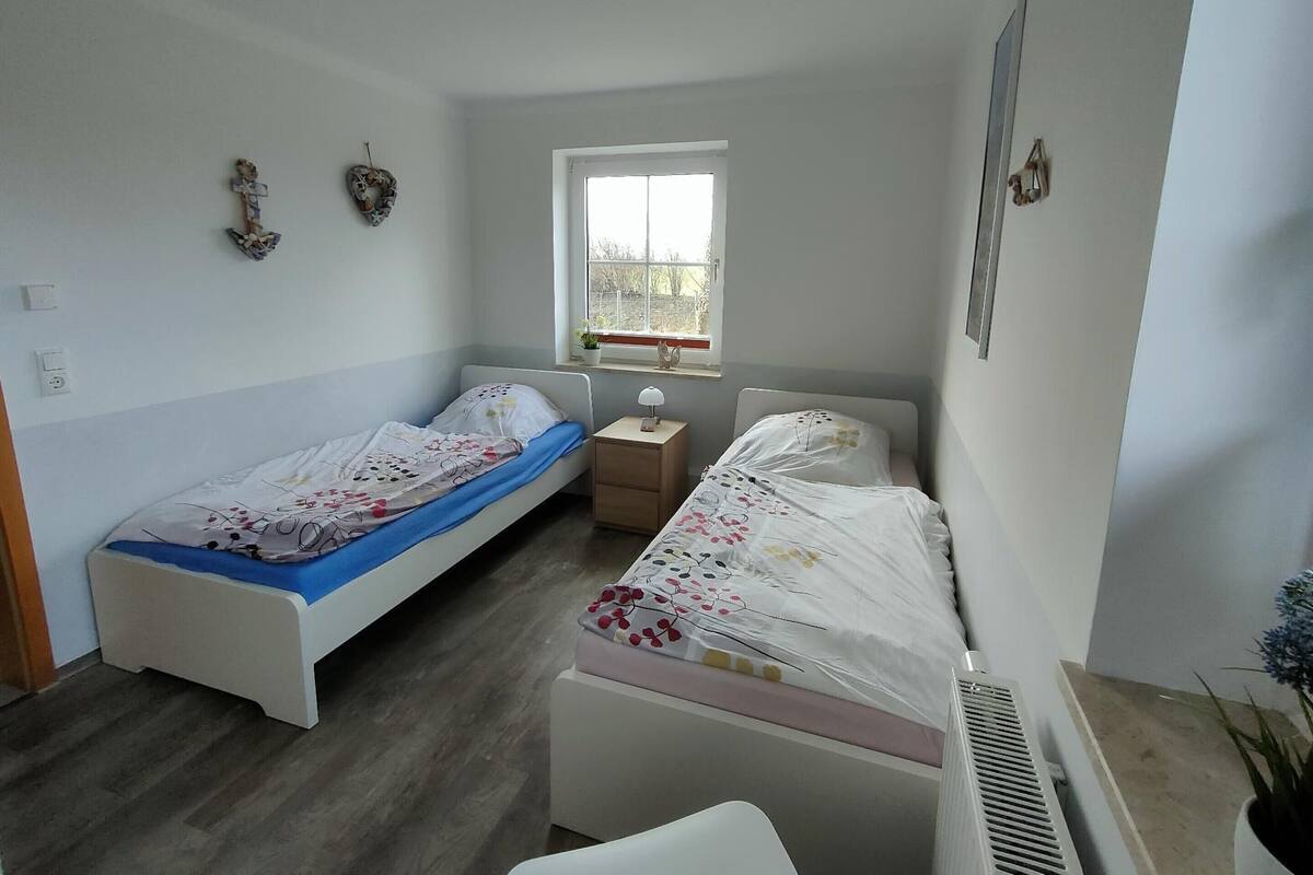 A small, well-lit bedroom with two single beds, a window, a bedside table, and a radiator.