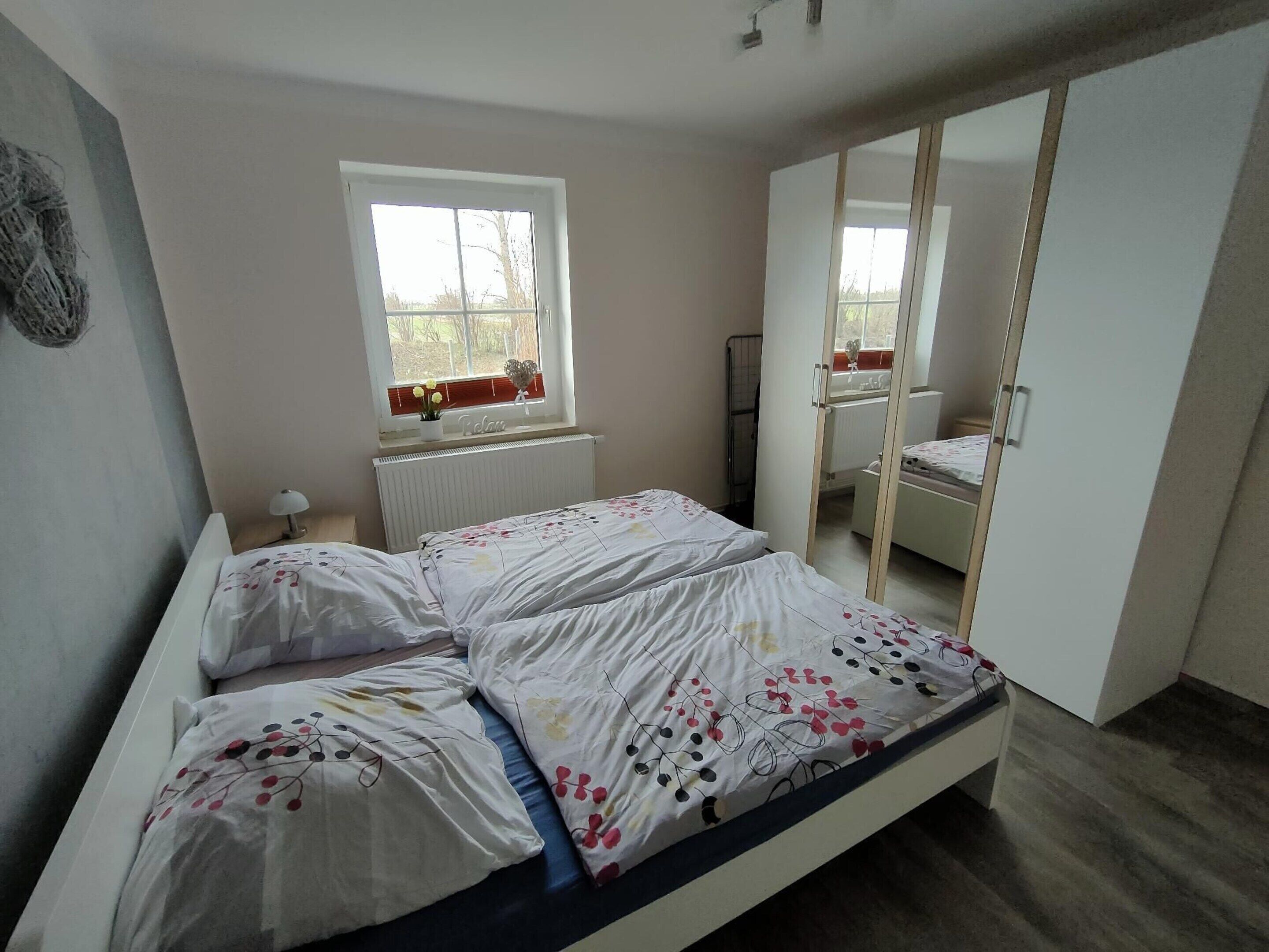 A bedroom with a bed, a window with flowers, a radiator, and a mirror.