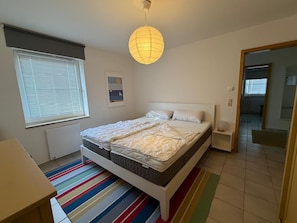 Ein Schlafzimmer mit einem Bett, einem Nachttisch, einem Fenster mit Jalousien, einem farbenfrohen StreifenTeppich und einer Pendelleuchte.