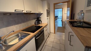 A modern kitchen with a stove, sink, and coffee maker. There is a view into the living area through an open doorway. - Holiday Complex Wenkendorf, Fehmarn-wenkendorf (Fehmarn)
