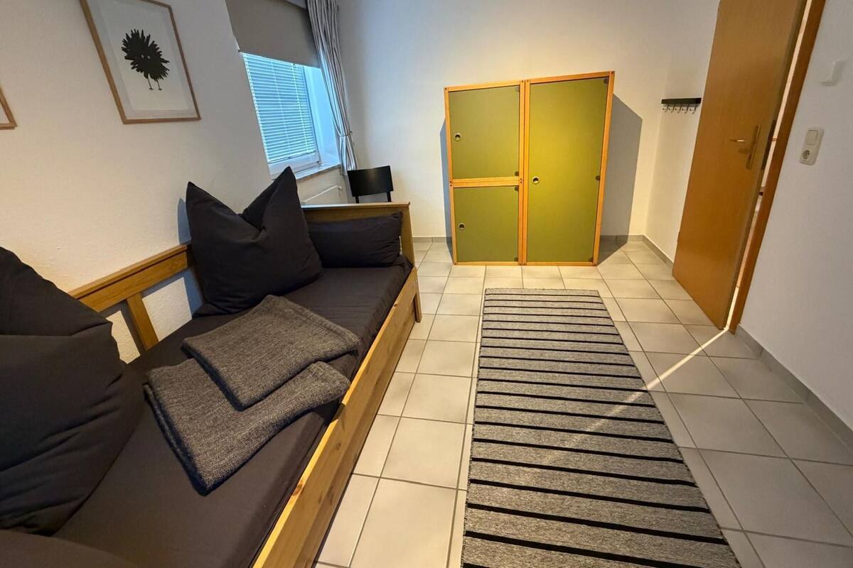 A room with a sofa, a wooden wardrobe, a window with blinds, and a tiled floor.