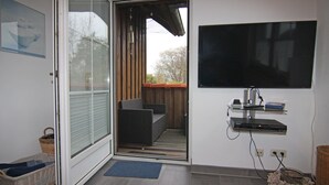 A room with a sliding glass door leading to an outdoor area with a wicker sofa and a TV mounted on the wall. - Holiday Complex Wenkendorf, Fehmarn-wenkendorf (Fehmarn)