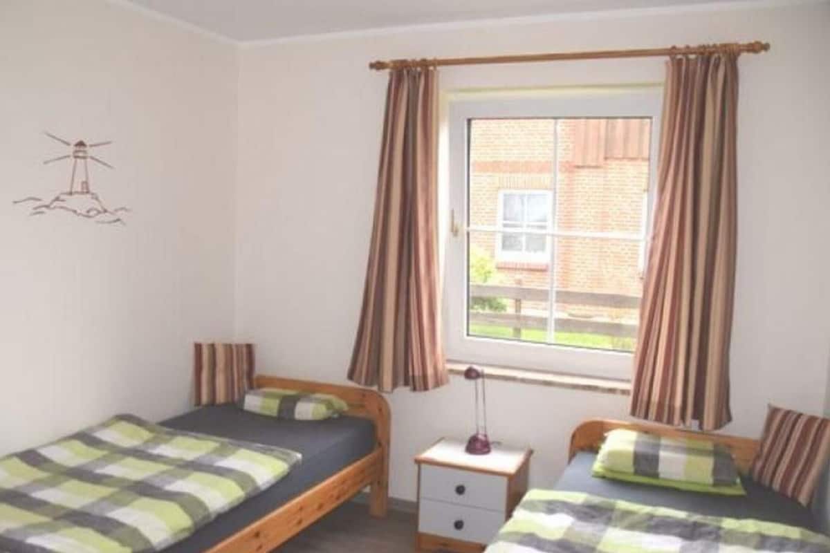 A bedroom with two single beds, a window with curtains, a bedside table with a lamp, and a wall decoration.