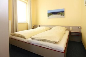 A single bed with white bedding in a room with yellow walls, a window with curtains, and a small bedside table. - Apartments Panorama With sea View Directly at the Beach Promenade of Laboe (Laboe)