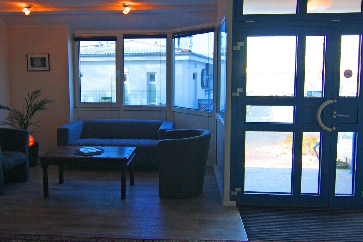 A living room with a sofa, a coffee table, and a potted plant. There is a large window with a view of buildings outside.