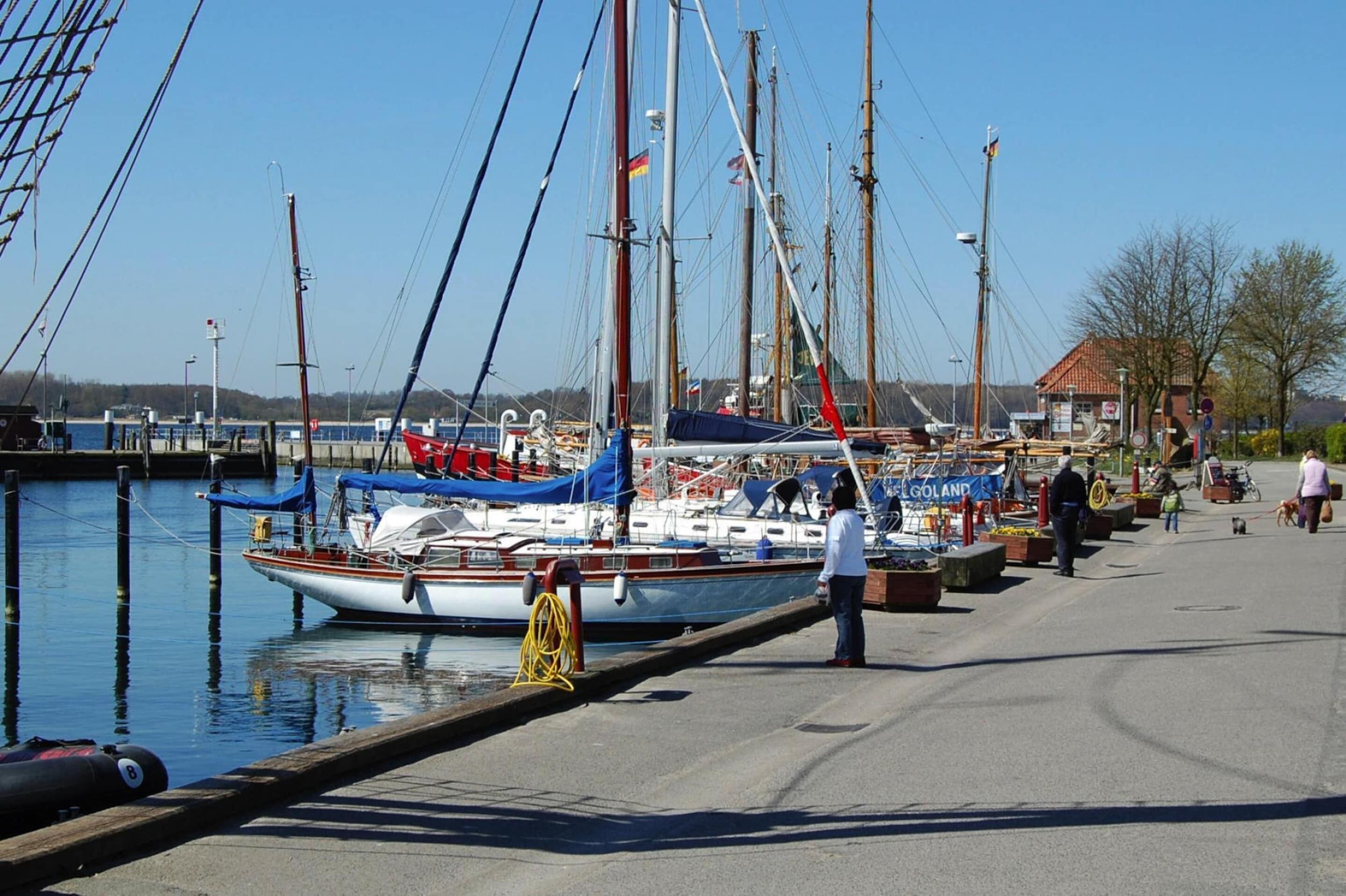Ein Yachthafen mit mehreren Segelbooten, einer Fußgängerpromenade und einem kleinen Gebäude mit Beschilderung.