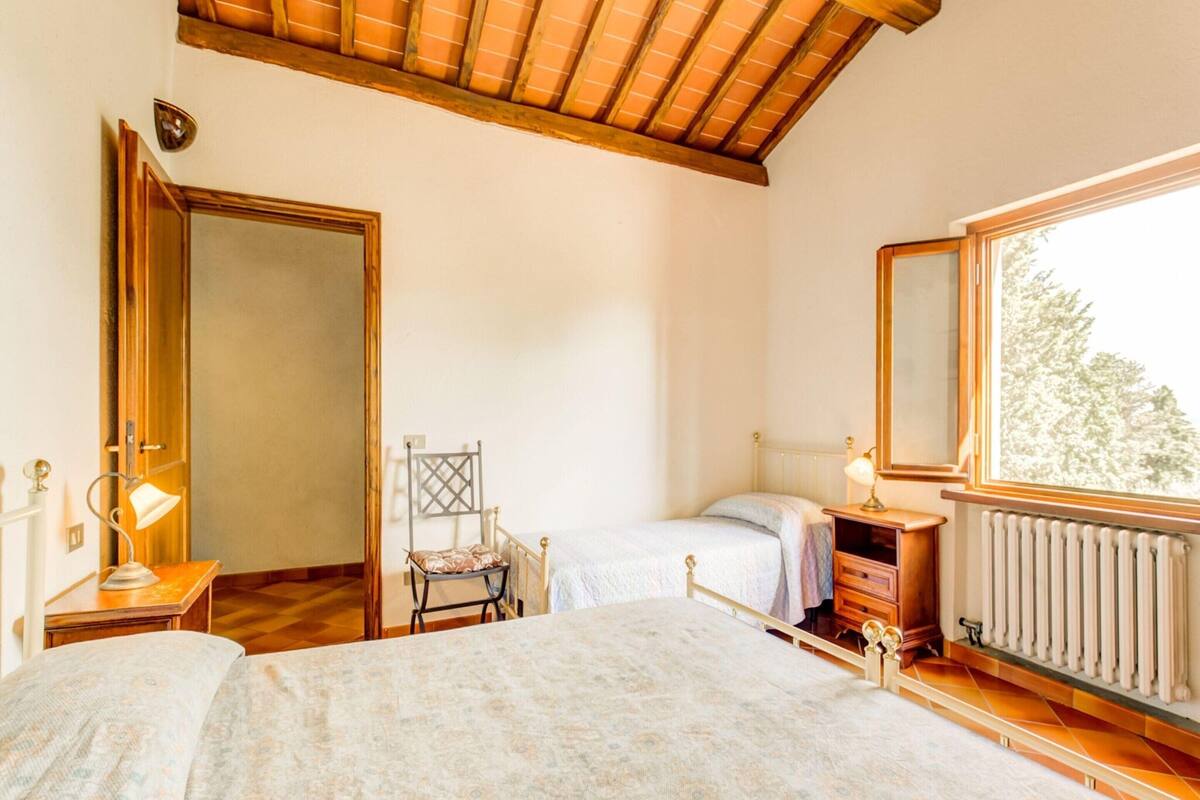 A bedroom with a wooden ceiling, a bed, a chair, a nightstand, a lamp, a dresser, and a window with a view of trees.
