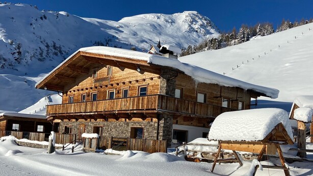 A wooden chalet with a stone facade, multiple windows, and a balcony, surrounded by snow-covered mountains. - Unique Large Alpine Pasture in the Middle of the Zillertal Mountains (Zellberg)