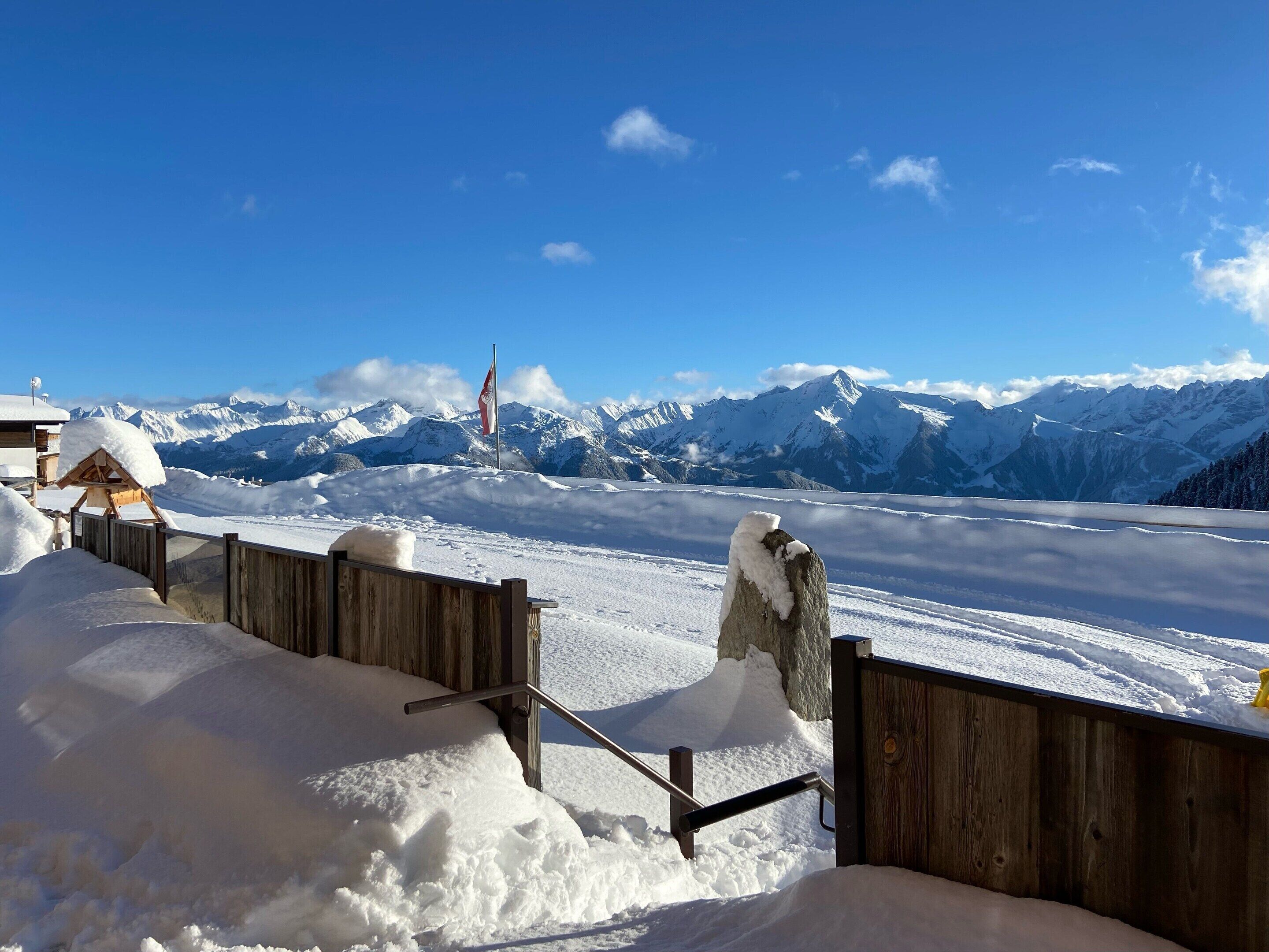 Una valla de madera con escalones cubiertos de nieve que conducen a un edificio, un asta con una bandera y una cordillera en el fondo.