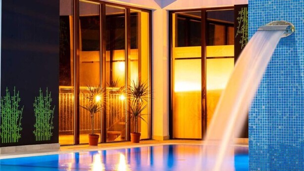 Una piscina interior con caída de agua, rodeada de puertas de cristal y plantas en macetas.