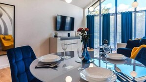 Un comedor moderno con una mesa redonda preparada para cuatro personas, un televisor de pantalla plana montado en la pared y un ventanal con cortinas azules.