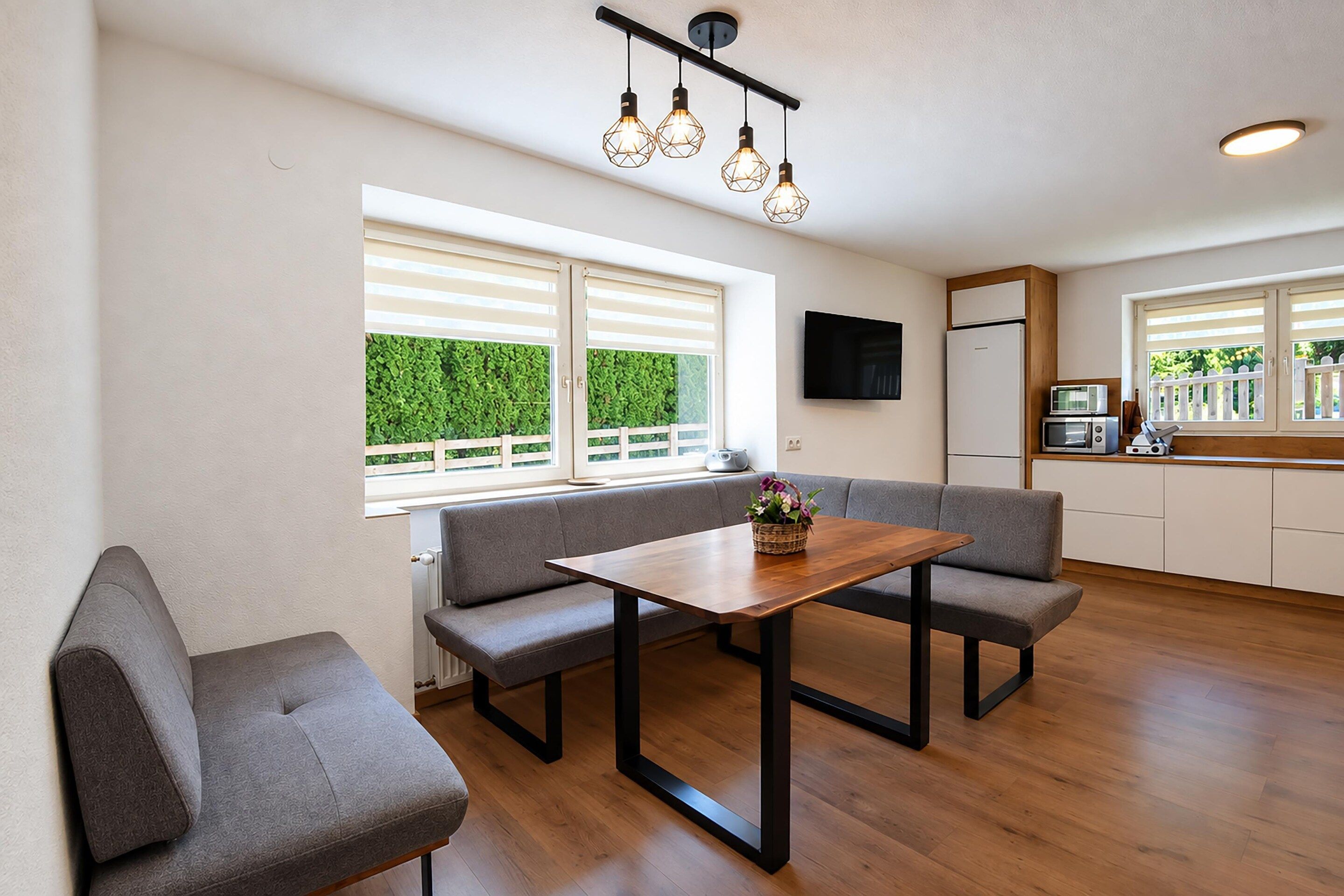 A modern kitchen with a dining area, wooden flooring, and a view of greenery outside.