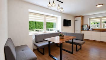 A modern kitchen with a dining area, wooden flooring, and a view of greenery outside.