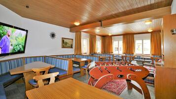 A cozy room with wooden benches and tables, a foosball table, a television, and large windows with curtains.