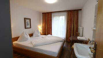 A hotel room with a wooden bed, a bedside table with a lamp, a bathroom with a sink and mirror, and a window with curtains.