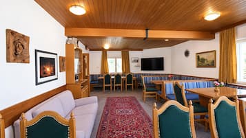 A cozy room with a sofa, a dining table, and a TV. There are wooden beams on the ceiling and a patterned rug on the floor.