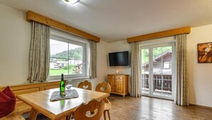 Ein Essbereich mit Holztisch, Stühlen und einer Bank. Ein Fernseher ist an der Wand befestigt, ein Fenster mit Vorhängen und Blick auf Gebäude.