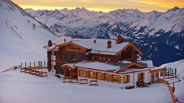 Eine mit Schnee bedeckte Berghütte mit Holzfassade und mehreren Fenstern, umgeben von verschneiten Bergen.