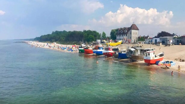 Une plage bordée de bateaux amarrés, un bâtiment doté d’une tour et, au loin, une zone boisée.