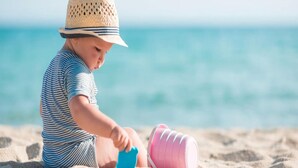 Ein Kind spielt mit Spielzeug an einem sandigen Strand, im Hintergrund ein klarer, blauer Himmel und ein Meer.