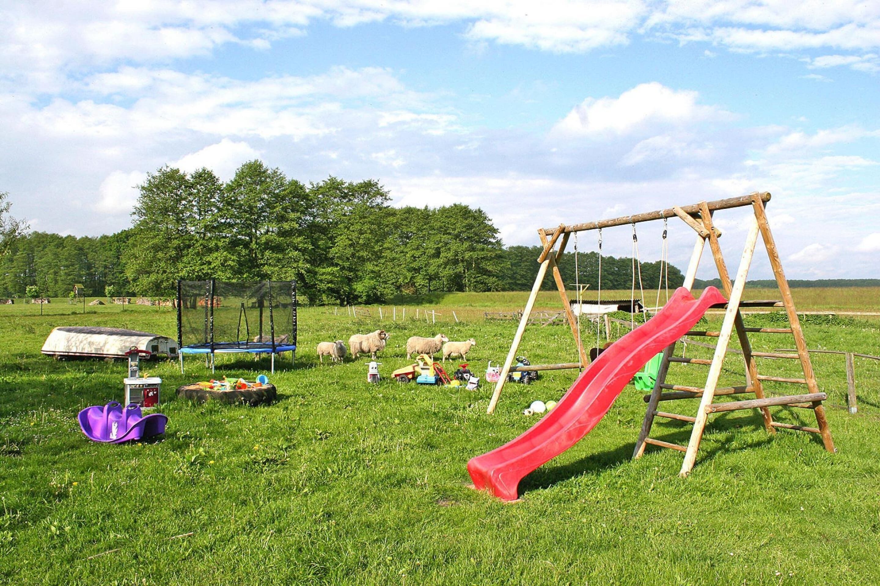 A playground with a wooden swing set and a red slide, surrounded by green grass and trees.