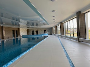 Ein großer, überdachter Swimmingpool mit klarem, blauem Wasser, einer langen Poollandschaft und großen Fenstern mit Blick ins Freie.