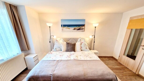 A neatly arranged bedroom with a bed, bedside lamps, a window with curtains, a radiator, and a wall-mounted picture.