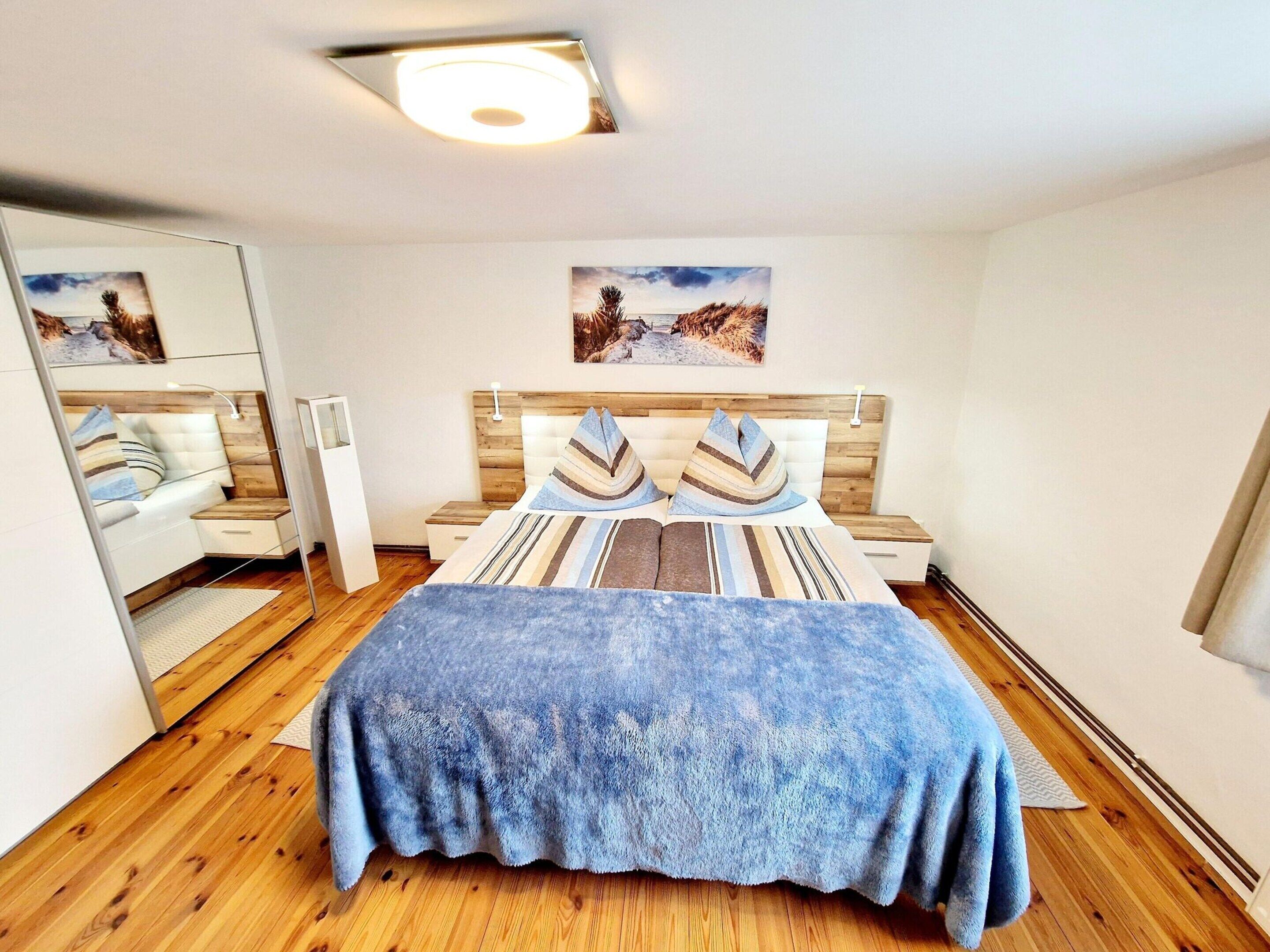 A bedroom with a wooden floor, a bed with striped bedding, a nightstand, a bedside table, a mirror, and a picture on the wall.