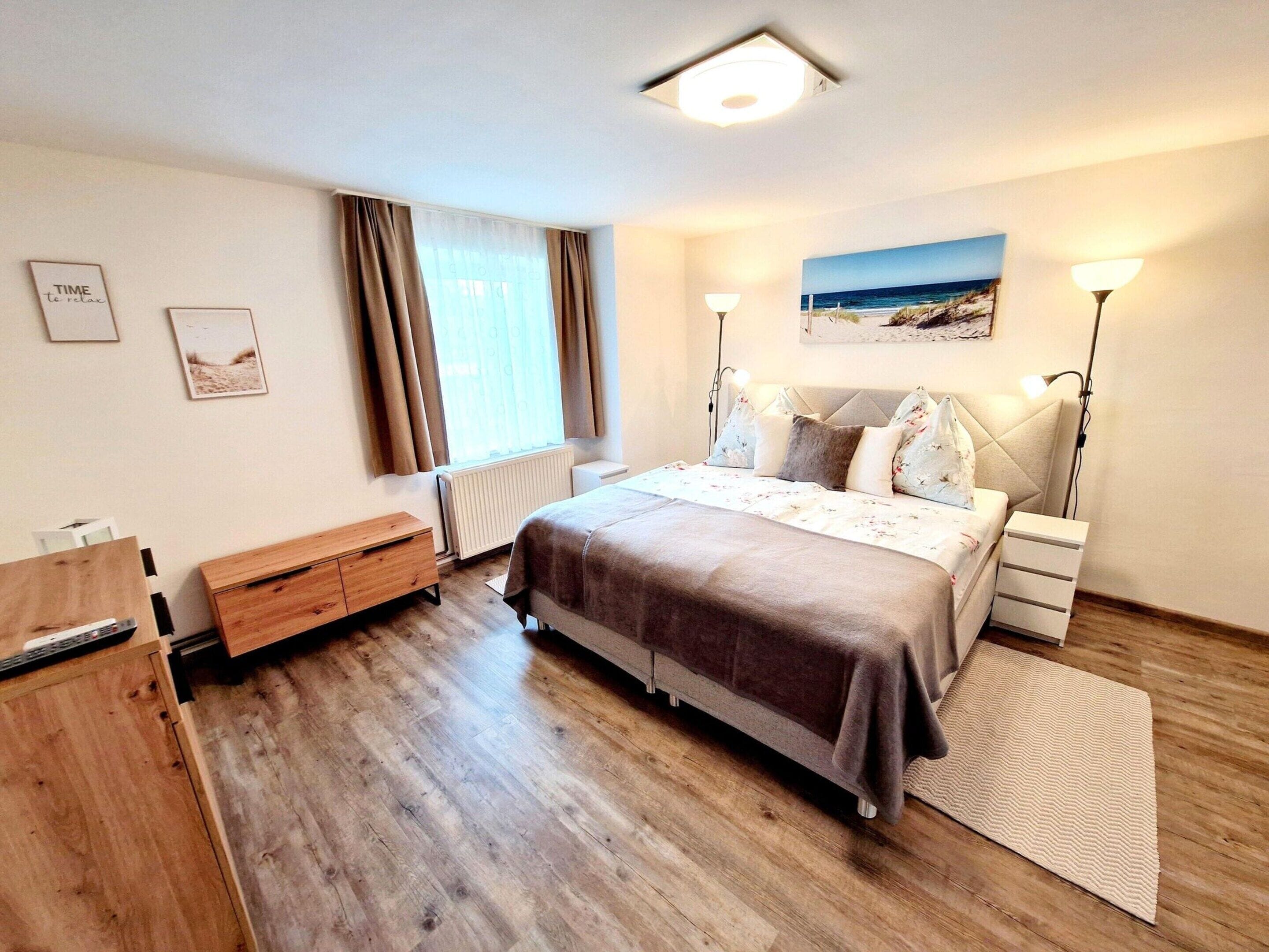 A modern bedroom with a bed, bedside lamps, a wooden dresser, a radiator, and a beach-themed painting on the wall.