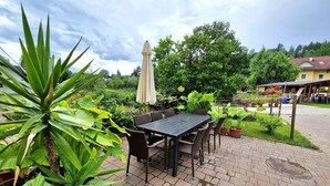 An outdoor patio with a table and chairs, surrounded by lush greenery and a cloudy sky.