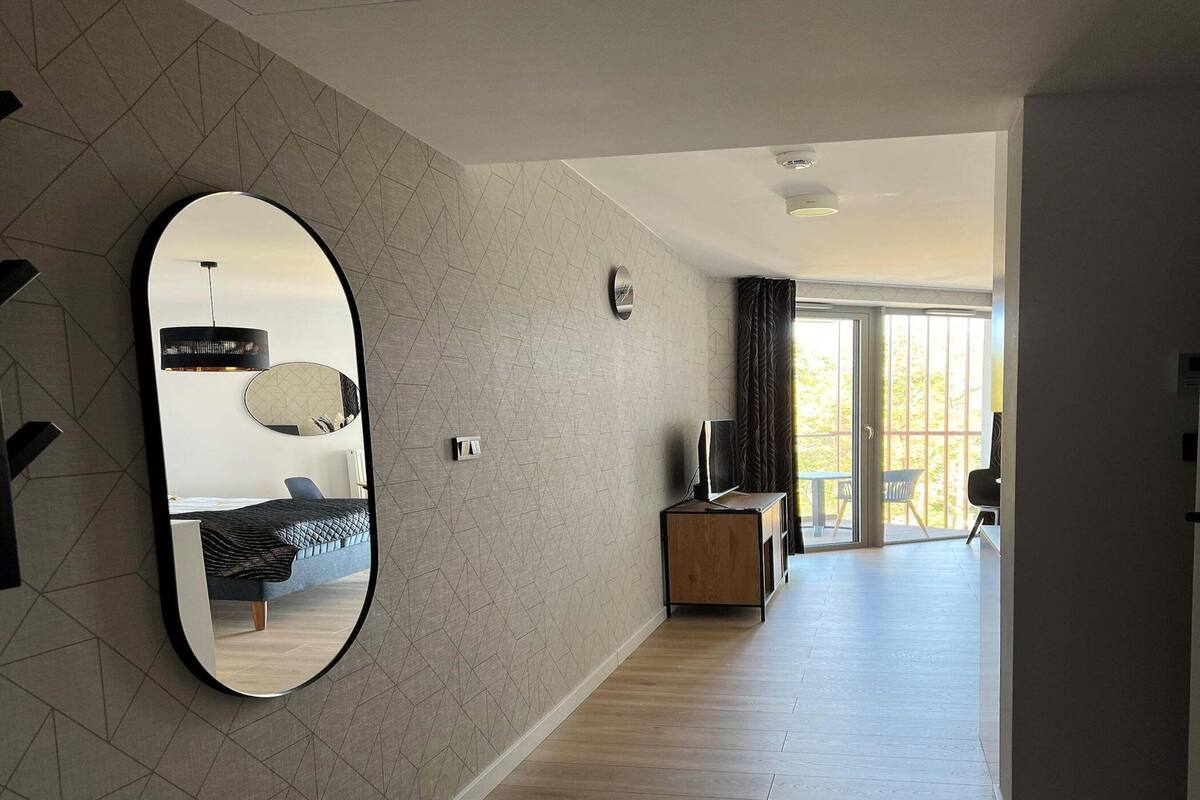 A modern hotel room with a geometric-patterned wall, a large mirror, a bed, a TV on a wooden stand, and a balcony with a view.