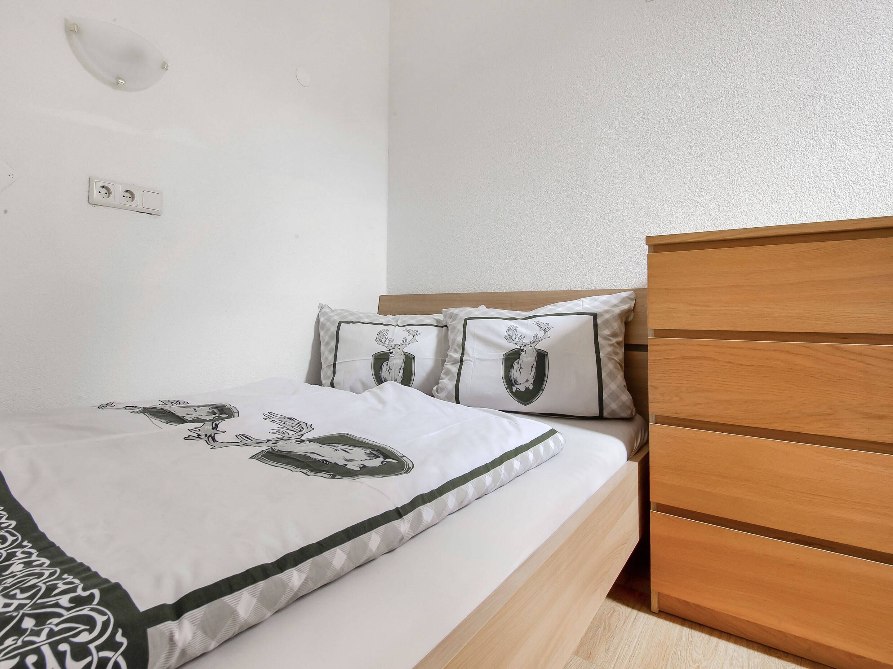 Een slaapkamer met een houten bedframe, een wit sprei met een grafisch ontwerp, twee kussens met een wapendiensign en een wandlamp, plus een stopcontact.