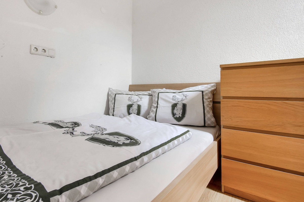 Ein Schlafzimmer mit einem Bettgestell aus Holz, einer weißen Bettdecke mit grafischem Muster, zwei Kissen mit Wappen und einer wandmontierten Leuchte sowie einer Steckdose.