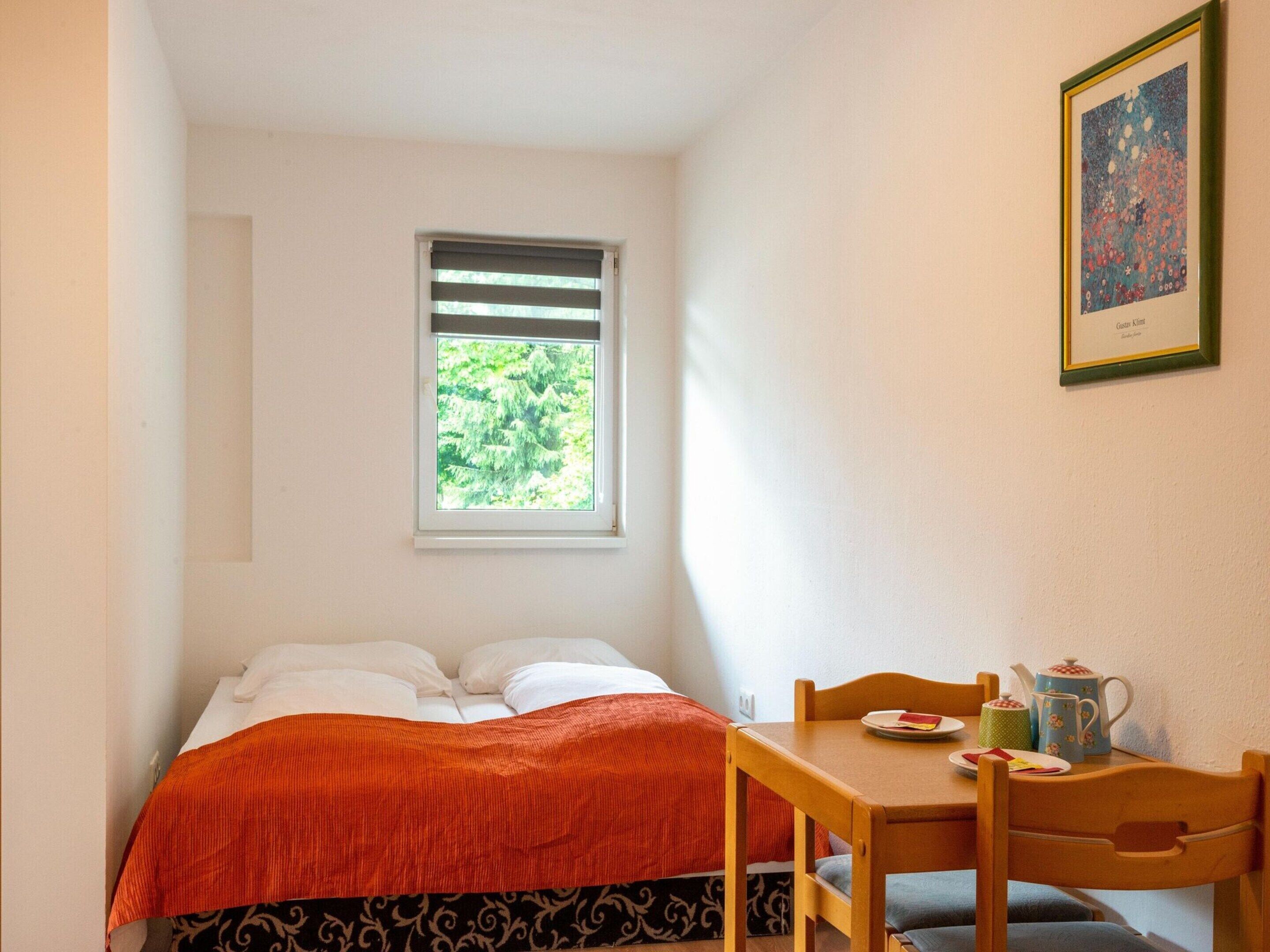 Ein kleines, ordentlich eingerichtetes Schlafzimmer mit Bett, Fenster mit Jalousien, Tisch mit Stühlen und einem gerahmten Bild an der Wand.