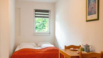 Ein kleines, ordentlich eingerichtetes Schlafzimmer mit Bett, Fenster mit Jalousien, Tisch mit Stühlen und einem gerahmten Bild an der Wand.