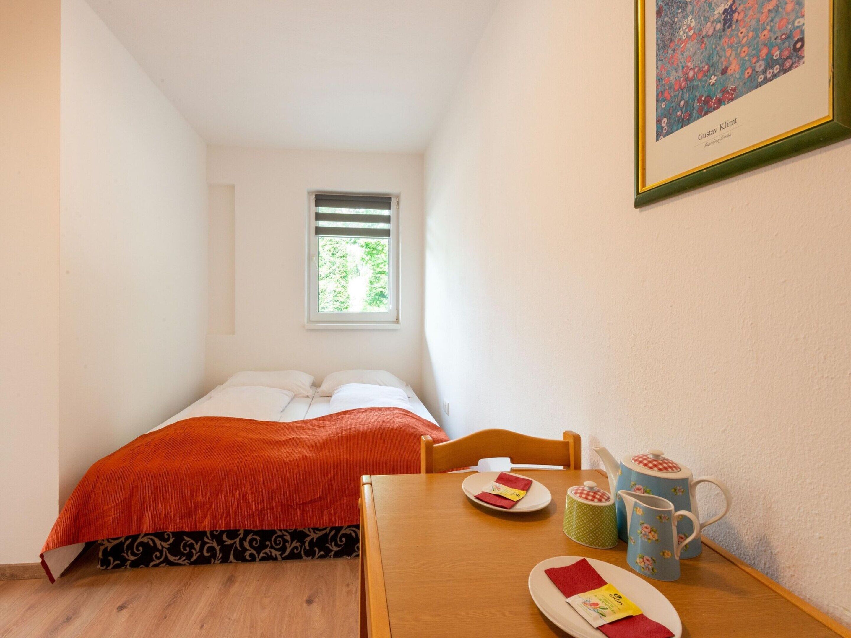 Une petite pièce chaleureuse, avec un lit simple, une table en bois sur laquelle se trouvent une tasse et une assiette, et un tableau encadré accroché au mur.