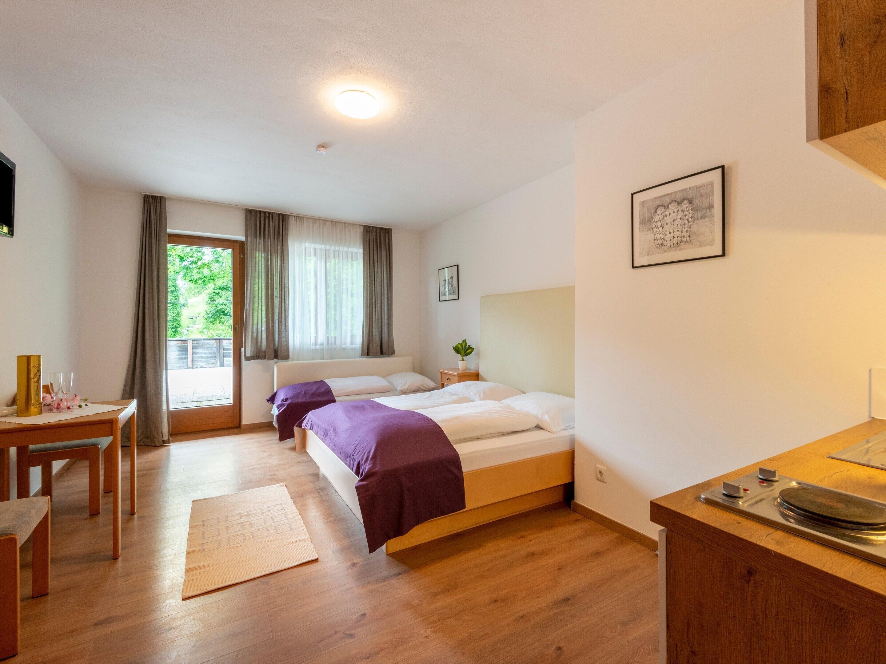 A hotel room with two beds, a wooden desk, a small table with a plant, and a window with curtains.