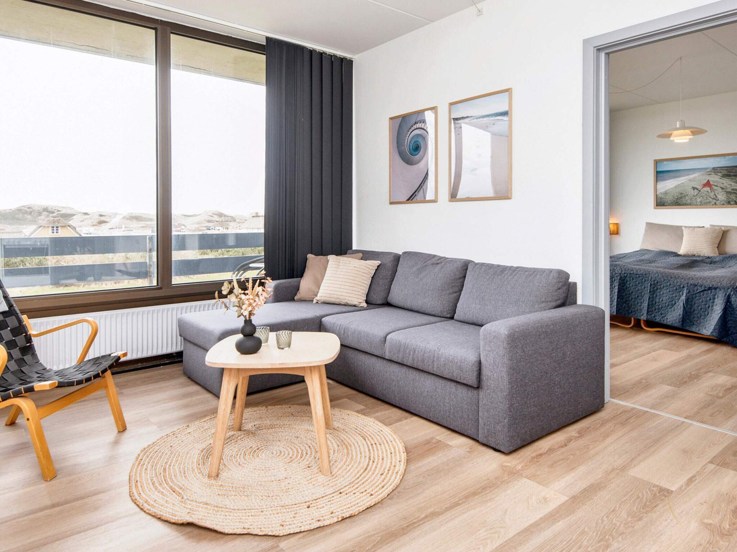 A modern living room with a grey sofa, a round wooden coffee table, and a woven rug. There is a view of the outdoors through large windows, and a bed with a blue blanket is visible in the adjacent room.