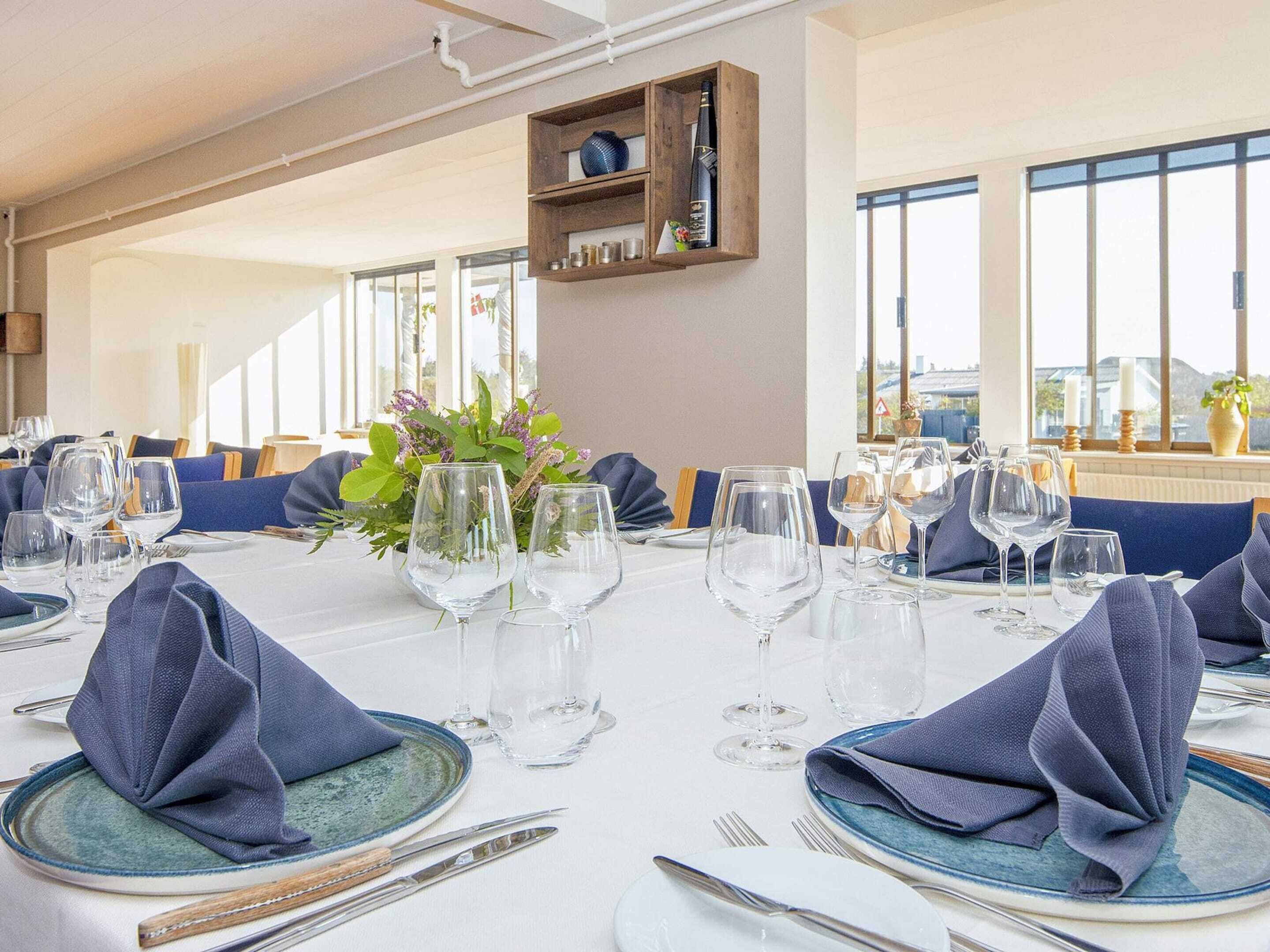 A dining area with tables set for a meal, featuring blue napkins, clear glassware, and a vase with flowers.