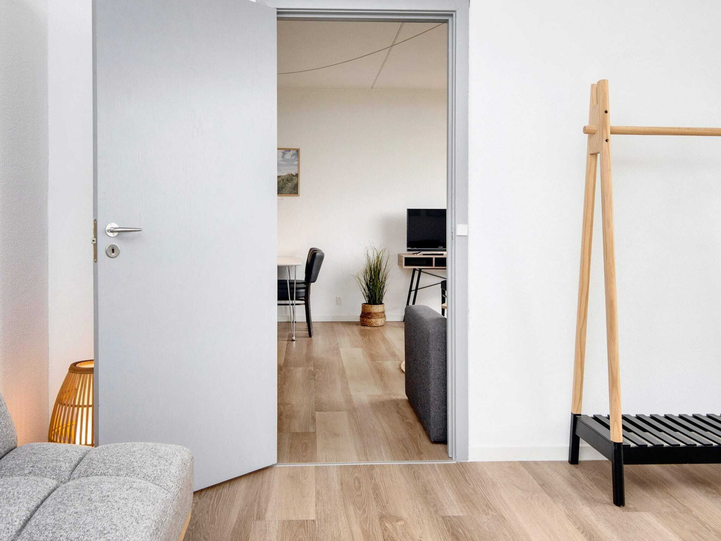A modern room with a wooden floor, a grey sofa, a black bench, and a door leading to a dining area with a table and chairs.